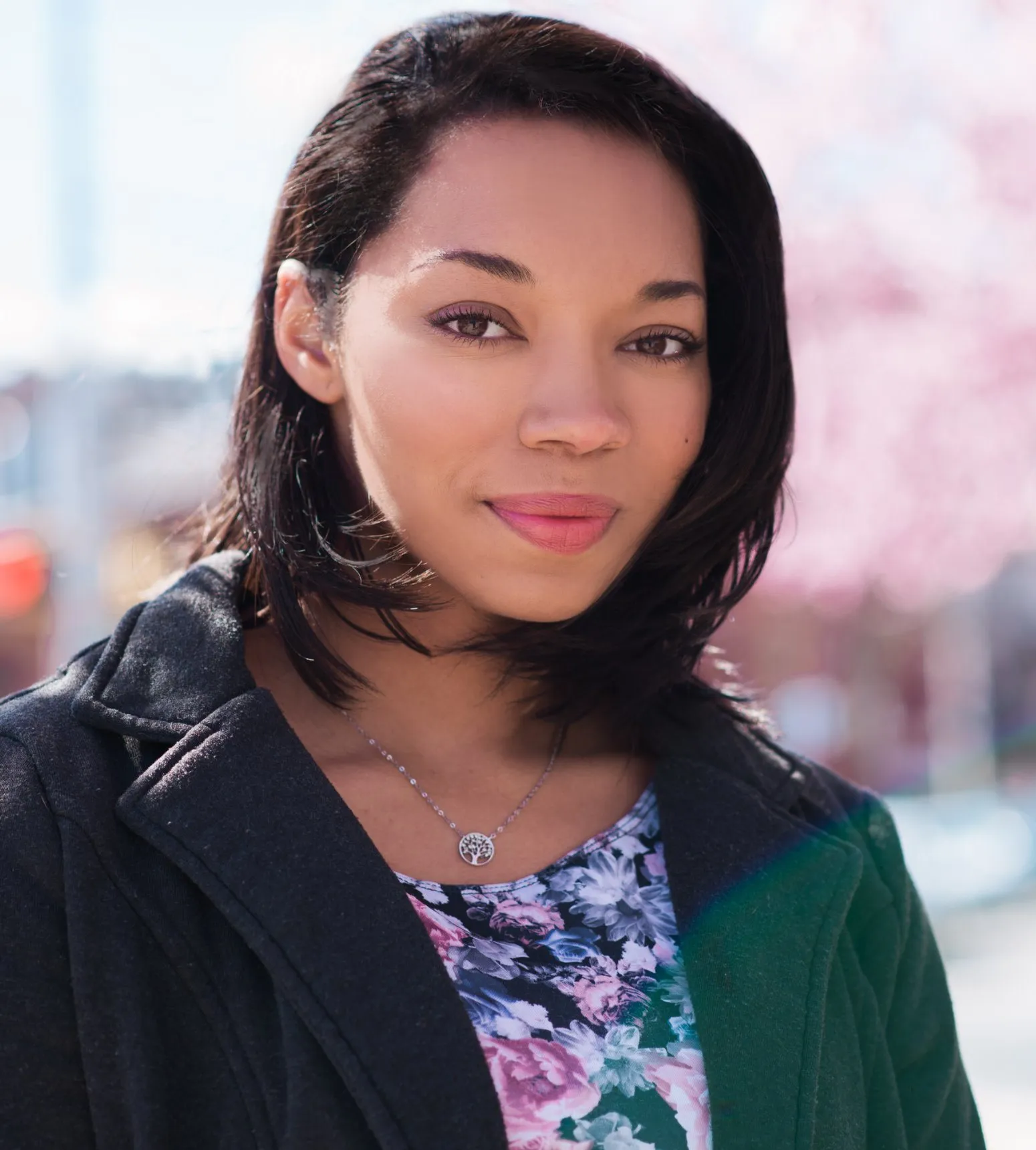 Alysa Buchanan outdoors in a dark coat over a floral top, smiling, with blossoming trees behind her.